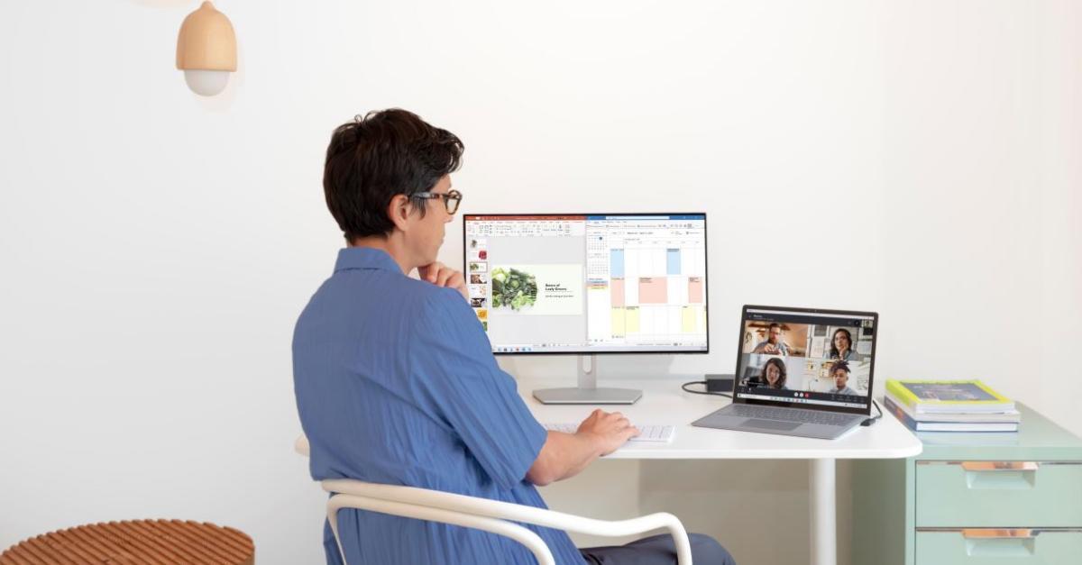 A woman sitting down having an online meeting on her laptop, with a computer screen and a wireless keyboard in front of her.