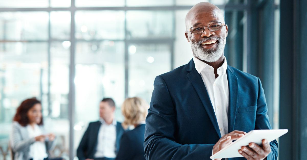 A man standing smiling and holding a tablet, with 3 coworkers sitting behind him.