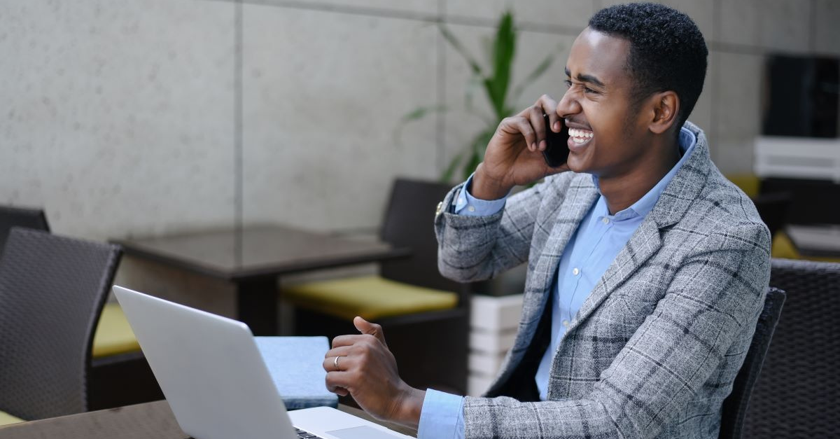 A man smiling talking on his mobile phone with a laptop in front of him.