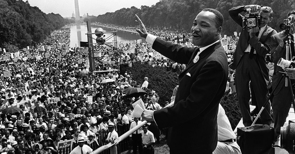 Martin Luther King Jr raising his right hand smiling, addressing a huge crowd.