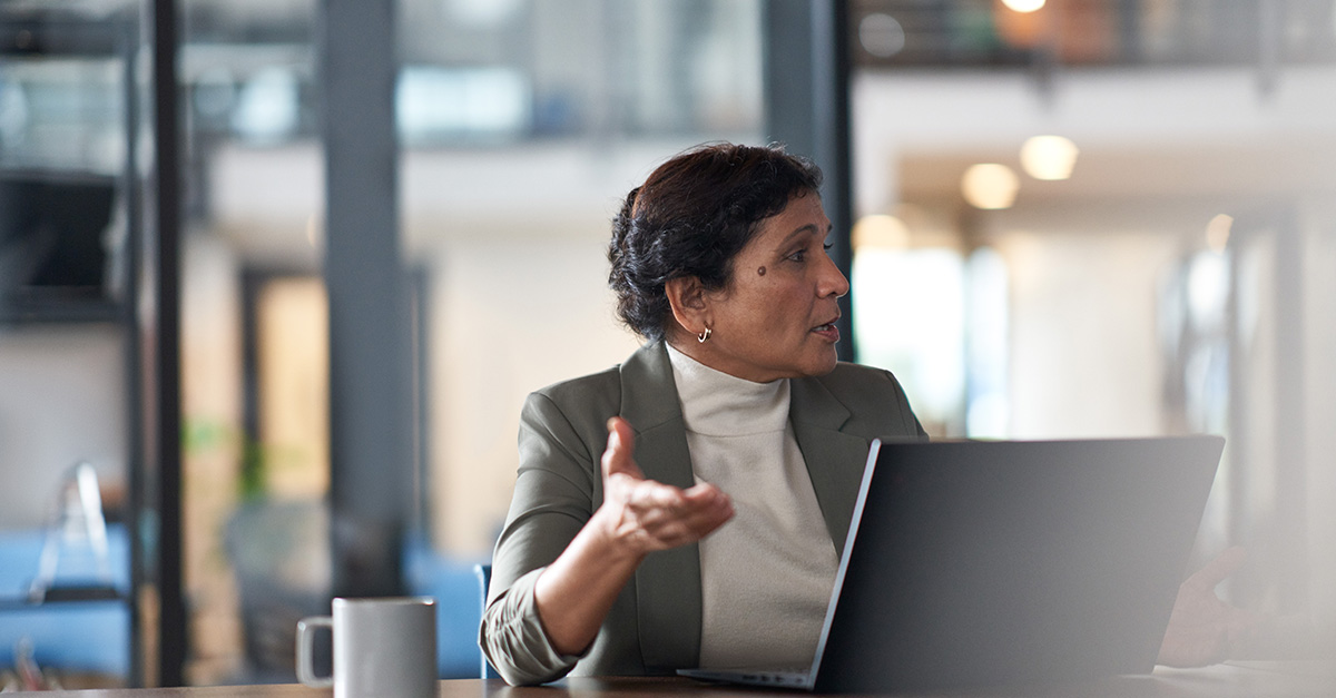 A woman working on her laptop, looking to her left while raising her right hand.