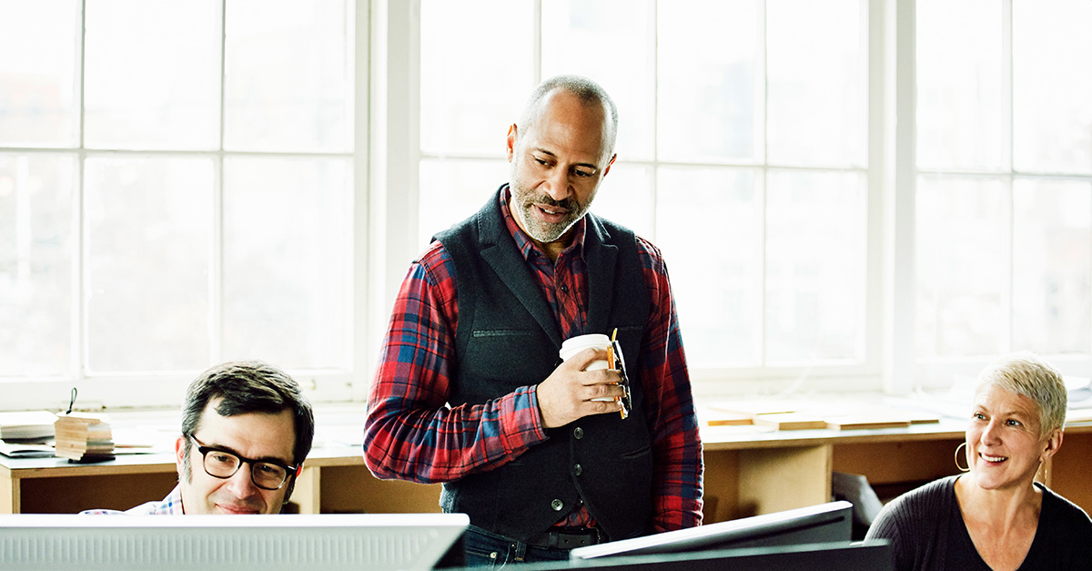 A man standing holding a cup of coffee, having a meeting with two fellow coworkers