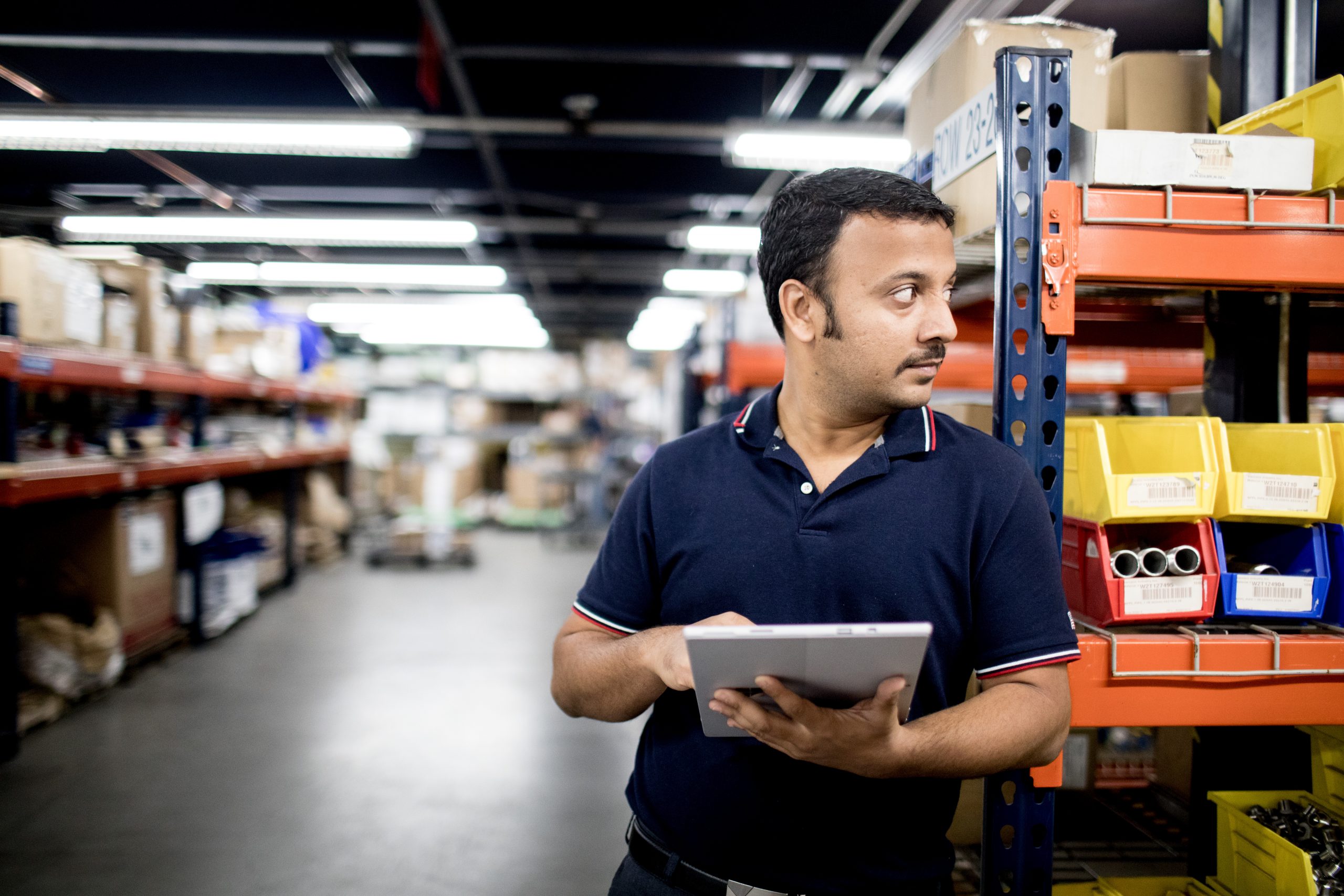 Factory worker standing in manufacturing warehouse among rows of metal shelving, holding a tablet.