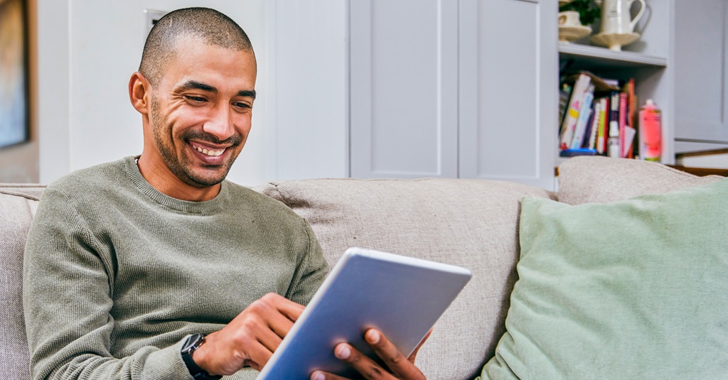 A smiling man sitting on a sofa looking at his tablet.