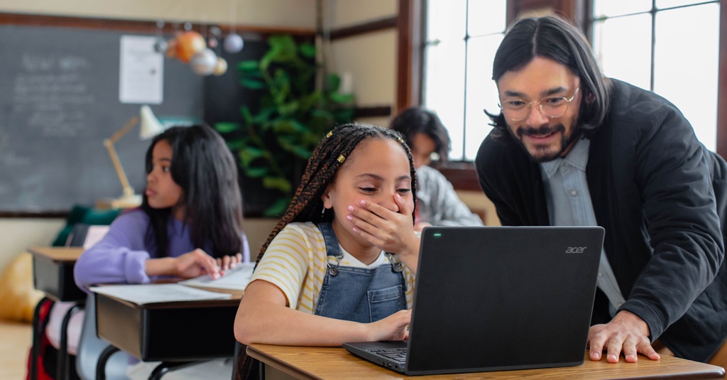 A teacher helps a student at her desk in a classroom.