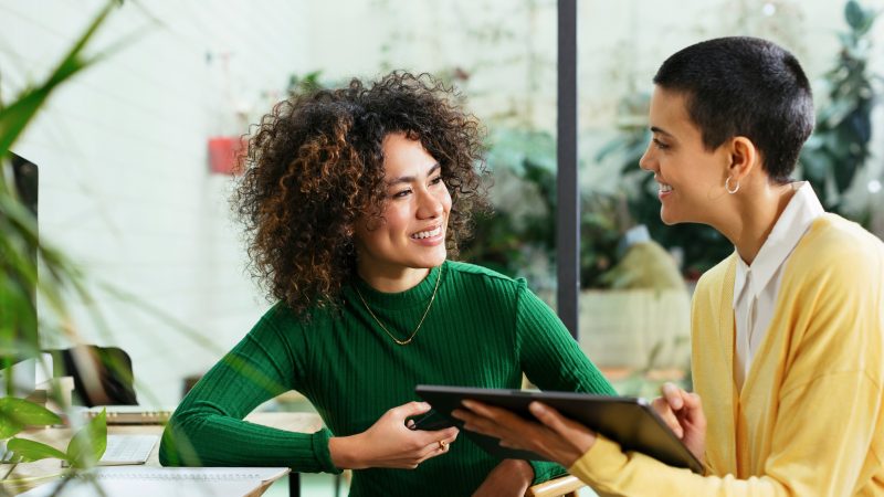 a person in a green shirt, talking with a person in a yellow shirt and holding a digital tablet together.