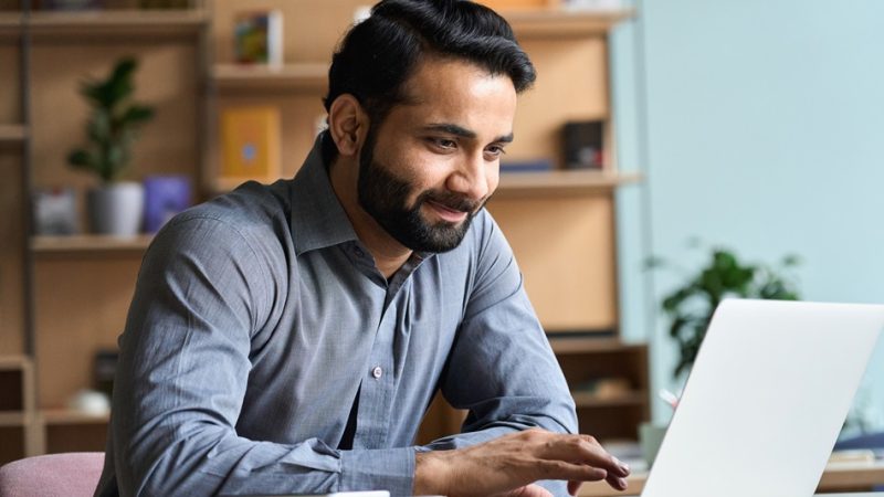 Un hombre en su escritorio, trabajando en una computadora portátil y sonriendo.