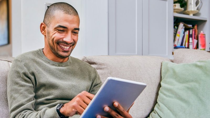 Un homme souriant assis sur un canapé, à l’aide d’une tablette..