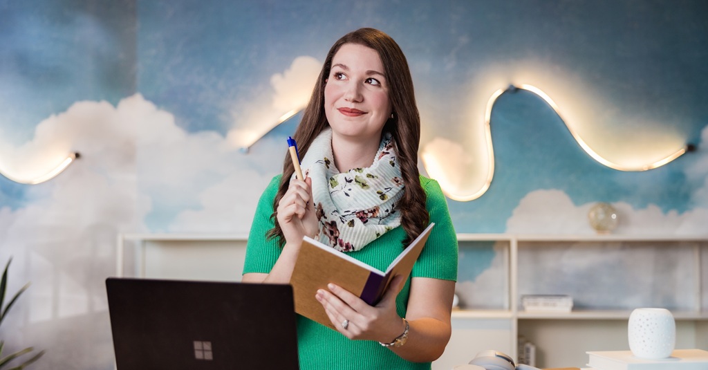 Femme tenant un stylo et un cahier, dans une pièce avec des nuages et un ciel bleu peints sur le mur.